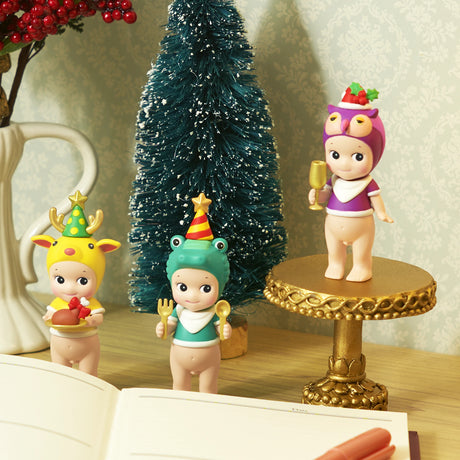 Three festive figurines from the Sonny Angel Christmas Dinner Blind Box, dressed in animal-themed costumes, stand near a book and a small tree.