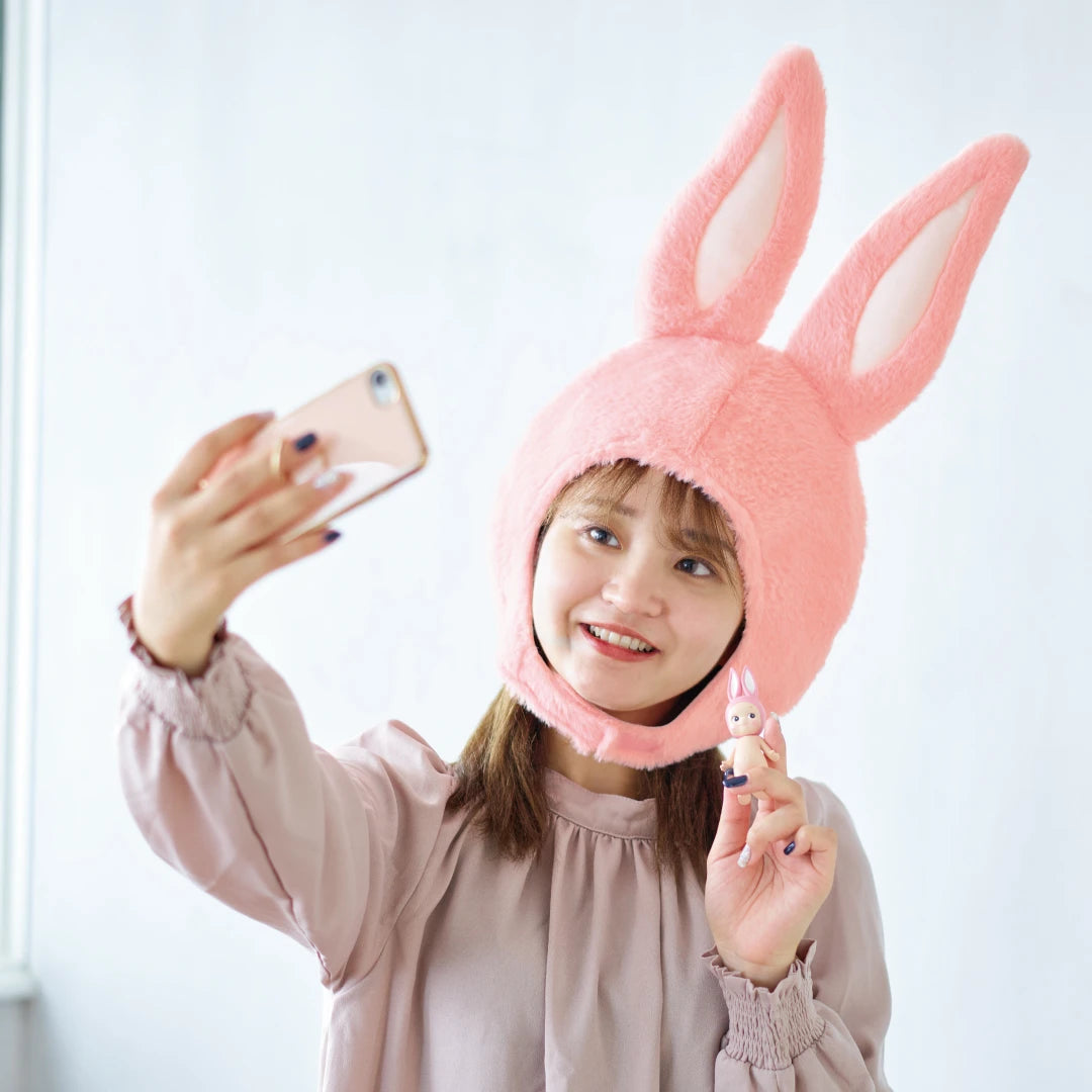 A person wearing a pink bunny hat takes a selfie while holding a small bunny figurine from the Sonny Angel Animal Costume - Series 2 collection by Sonny Angel.