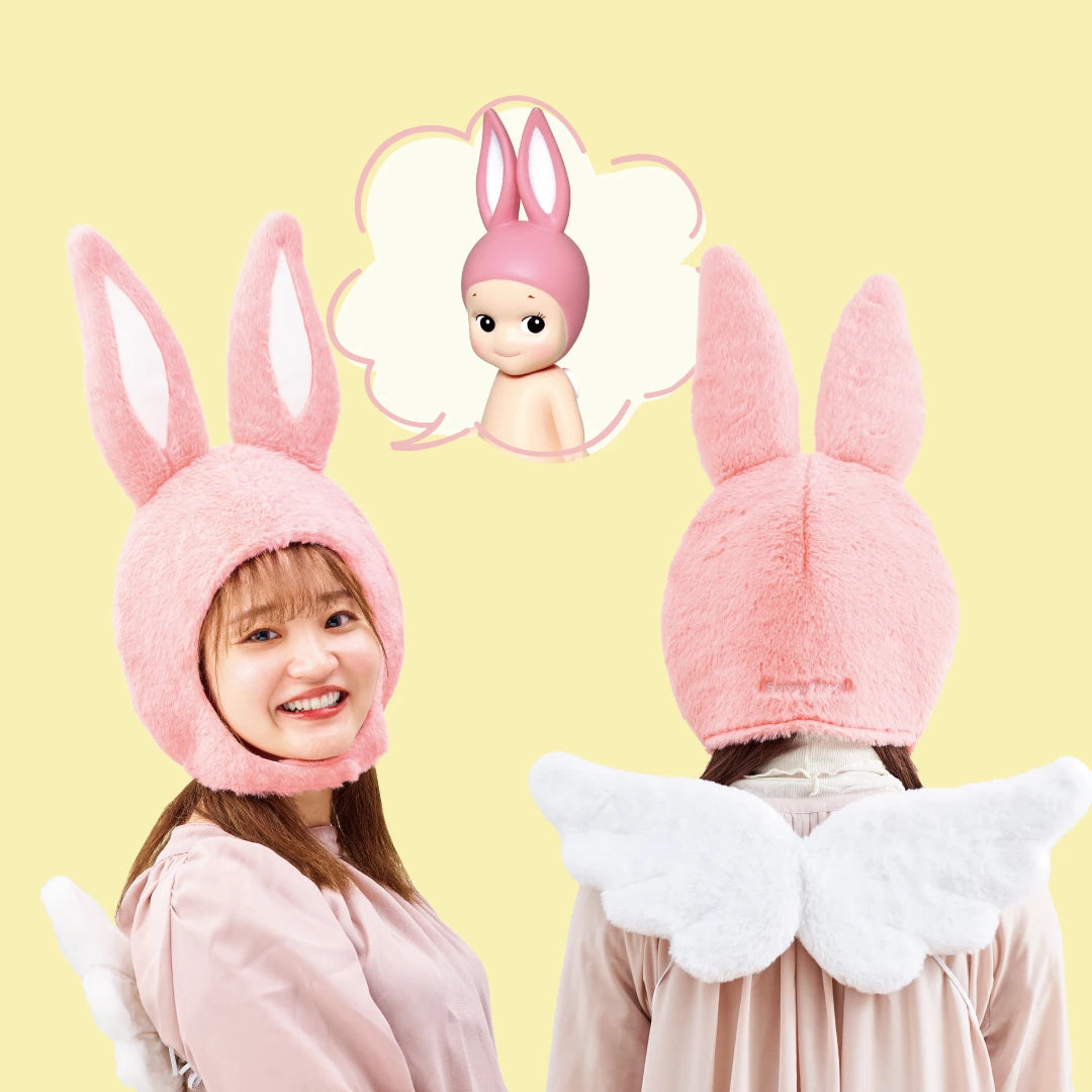 Two individuals are wearing pink bunny hats and white wings. The person on the left is smiling at the camera, while the other is facing away. A thought bubble above them features a Sonny Angel from Sonny Angel Animal Costume - Series 2, also adorned with a delightful bunny hat.