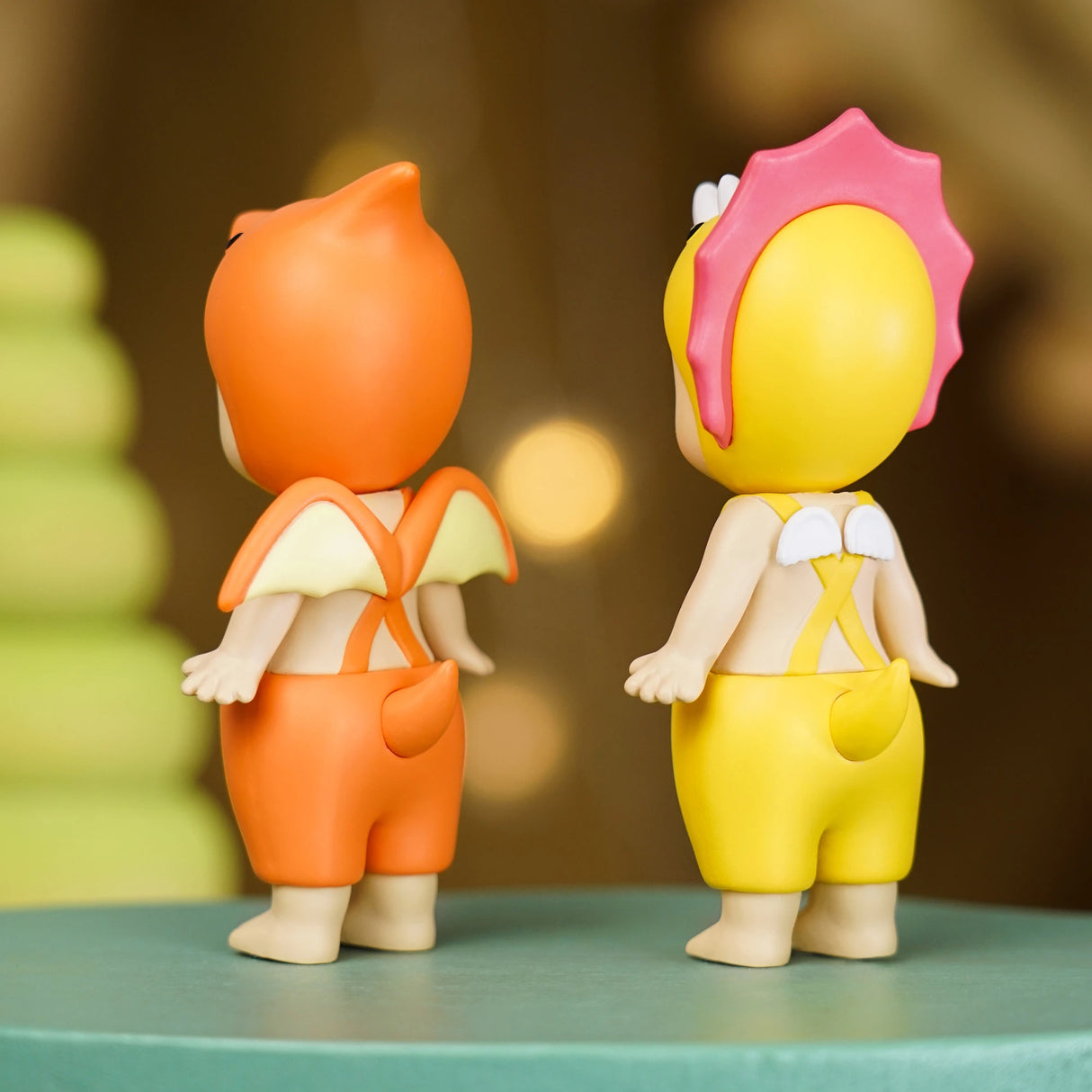 Two vibrant figures from the Sonny Angel Dinosaur Series, by the brand Sonny Angel, are displayed on the table. One figure is orange with bat-like wings, and the other is yellow with a pink headdress and small wings. Both figures are seen from behind, showcasing these charming prehistoric characters from this limited edition series.