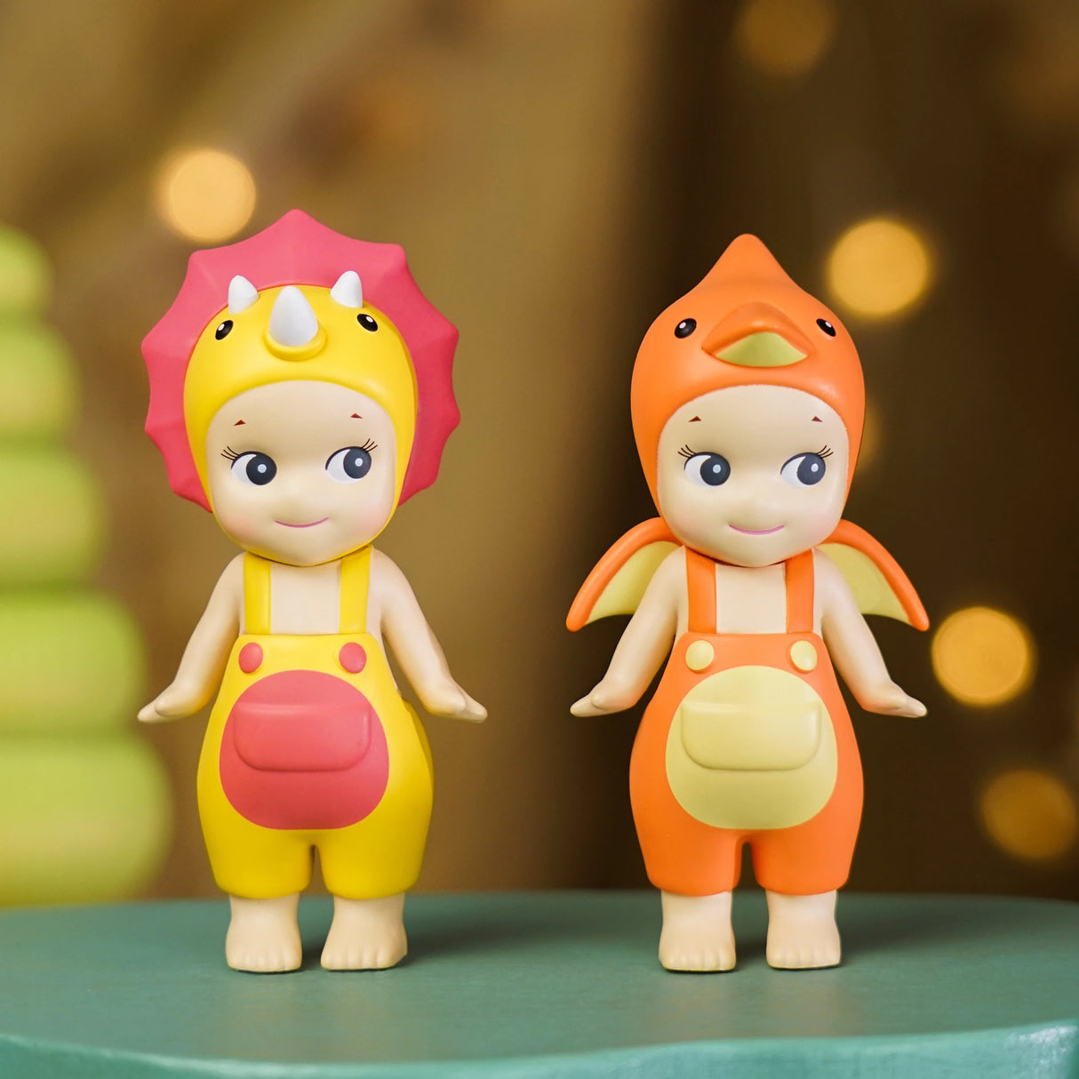 Two figures from the Sonny Angel Dinosaur Series by Sonny Angel are dressed in yellow and orange dinosaur-themed outfits, standing side by side on a green surface with a blurred background.