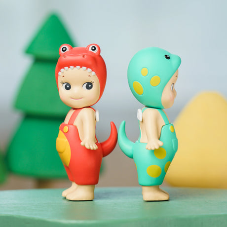 Two adorable figures from the Sonny Angel Dinosaur Series, one in red and the other in green, are positioned back-to-back on a surface. These charming prehistoric pals are set against a lively, colorful backdrop featuring trees.