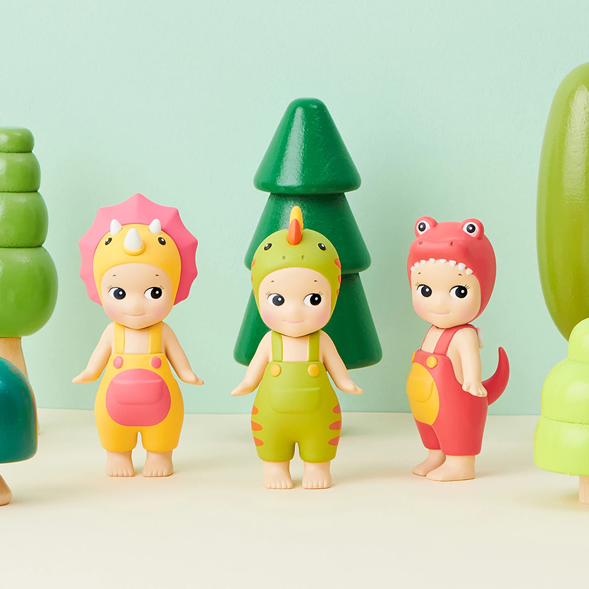 A trio of figures from the Sonny Angel Dinosaur Series by Sonny Angel, each adorned in dinosaur costumes, are positioned in front of playful cartoonish green trees set against a light green backdrop. These charming prehistoric companions add a touch of whimsy to any collection.