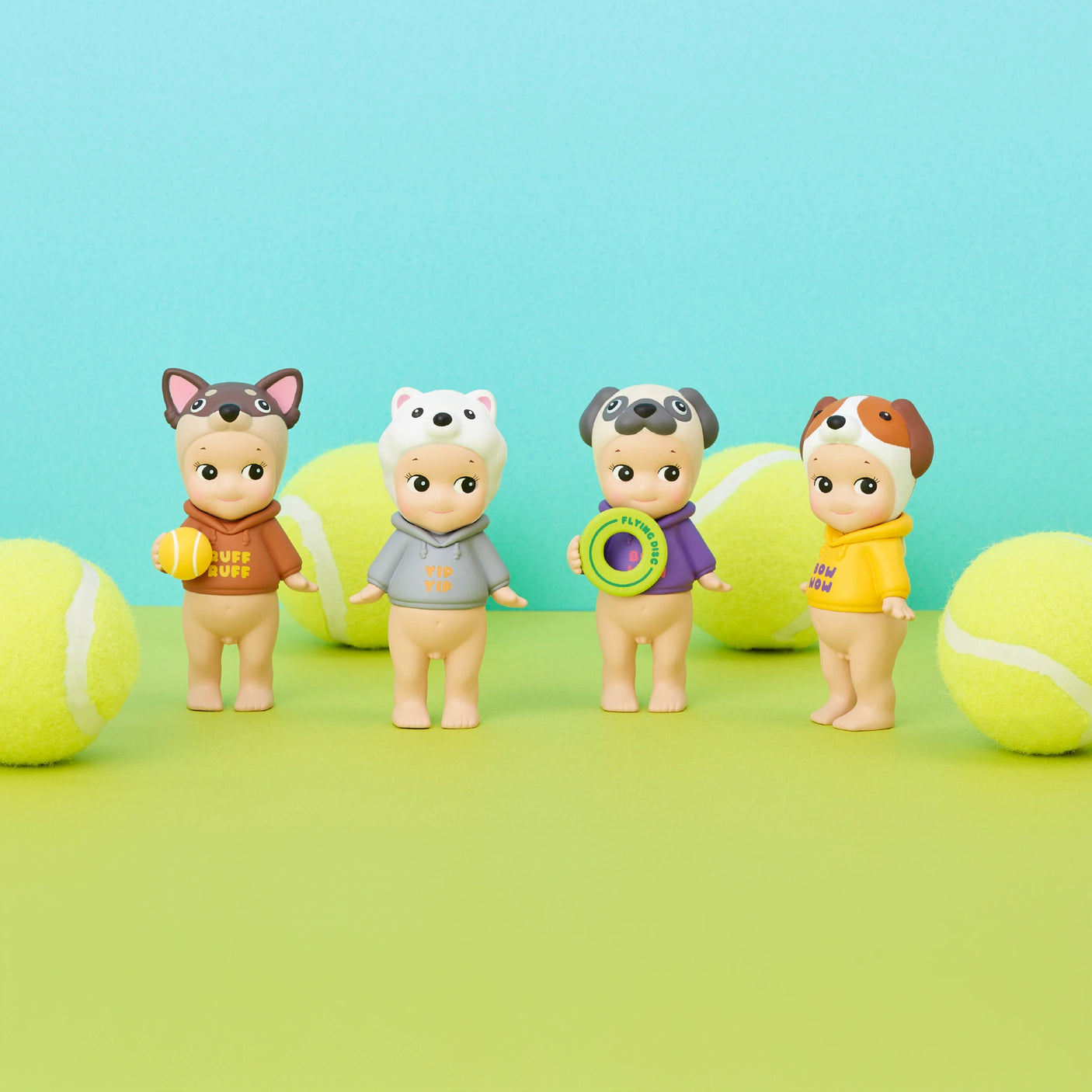 Four collectible figures from the Sonny Angel Dog Time Series, each dressed as a dog with a tennis ball, are showcased against a vibrant two-toned green and blue backdrop, perfectly capturing the playful essence of the series.