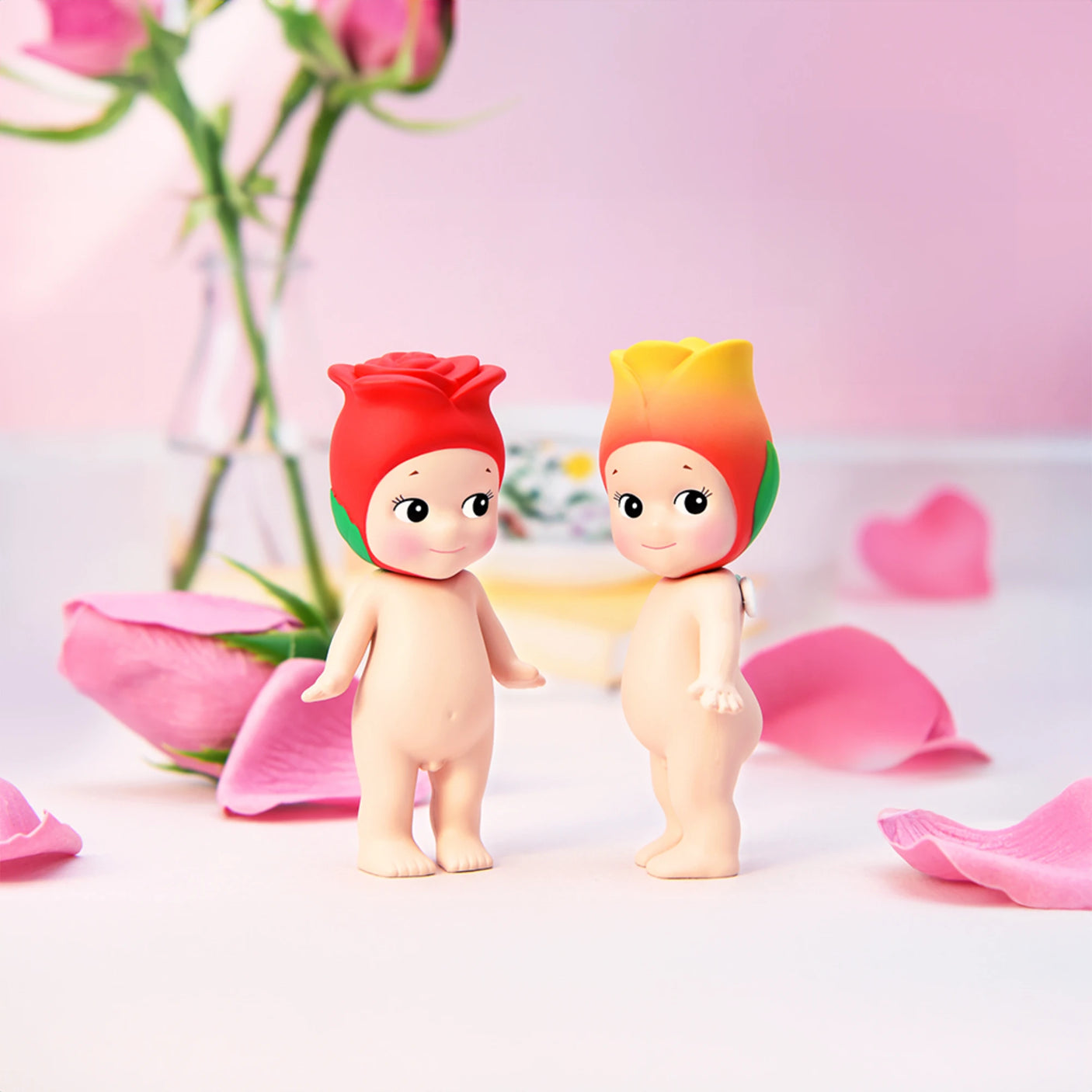 On a pink surface adorned with rose petals and set against a blurred floral background, two charming small figures from the Sonny Angel Flower Series stand out with their flower petal hats. These delightful toys from Sonny Angel capture the whimsy and surprise inherent in a blind box collection.