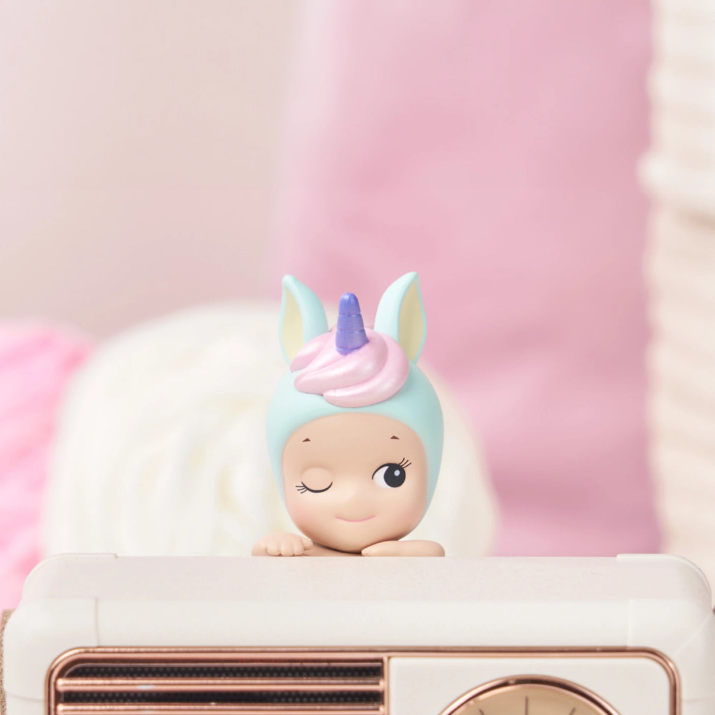 A small figurine from the Sonny Angel Hippers Blind Box - Dreaming series winks, its pastel unicorn horn adorning the hood as it peeks over a surface. The background is softly blurred in shades of pink and white, enhancing the whimsical allure of this dreamy Sonny Angel creation.