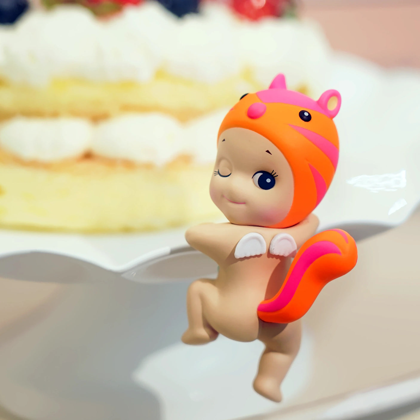 A small figurine from the Sonny Angel Hippers Blind Box - Looking Back series clings to a cake stand near a layered cake topped with whipped cream and berries, wearing a pink and orange squirrel hat. This delightful collectible from the Sonny Angel brand adds charm to any scene.