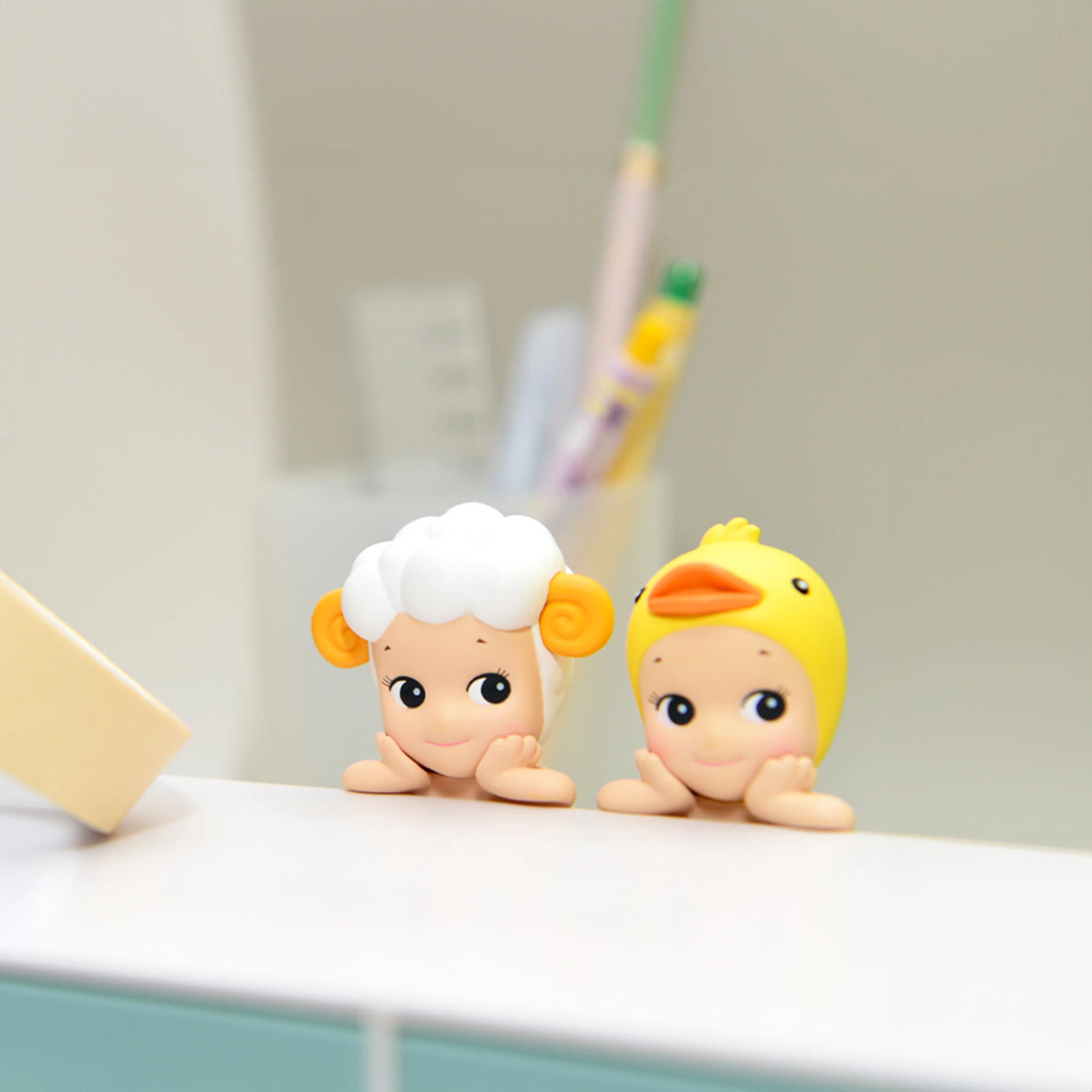 Two small, cartoonish figurines from the Sonny Angel Hippers Blind Box - Animals collection rest on a white surface. One figurine resembles a sheep with curly horns, while the other looks like a duck wearing a yellow beak and hat. In the background, blurred pens are held upright with reusable adhesive.