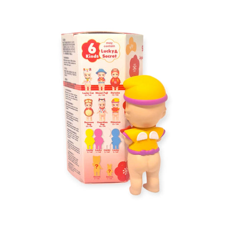 A Sonny Angel Japanese Good Luck figurine, dressed in yellow and purple, is positioned beside a packaging box that displays various toy designs inspired by traditional Japanese Lucky Charms.