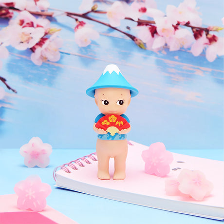 A petite figure, similar to a Sonny Angel Japanese Good Luck, wearing a blue hat is placed on a pink spiral notebook. Cherry blossoms surround the setting, creating an atmosphere of good fortune.