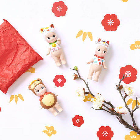 Three Sonny Angel Japanese Good Luck figures, dressed in animal-themed costumes, are arranged on a decorated backdrop featuring red flowers and a branch of white blossoms. This scene captures the spirit of traditional Japanese Lucky Charms and is complemented by a crumpled piece of red paper.