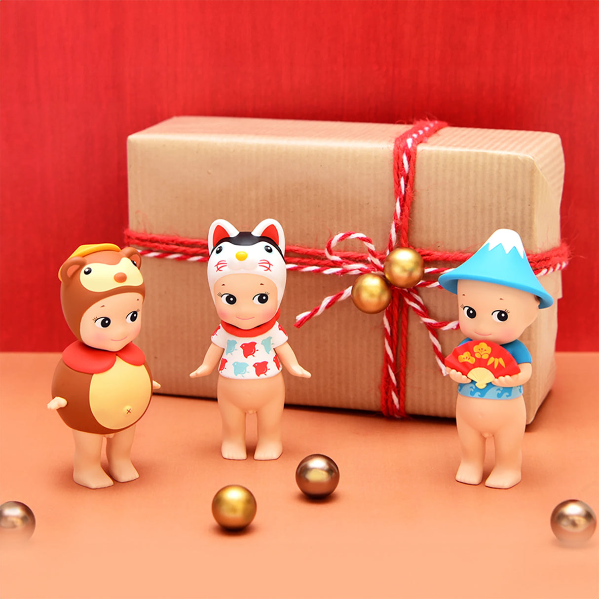 Three figures of Sonny Angel Japanese Good Luck toys, dressed in animal and mountain-themed costumes, are positioned before a gift box wrapped with a red ribbon. They are surrounded by gold spheres on a vibrant red background, capturing the essence of Japanese Lucky Charms.