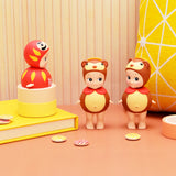 Three figures from the Sonny Angel Japanese Good Luck collection are showcased on a table set against a yellow and orange striped background. Two Sonny Angel figures, adorned in bear costumes, symbolize good fortune, while a red doll reminiscent of Japanese lucky charms is perched on a cylindrical stand.