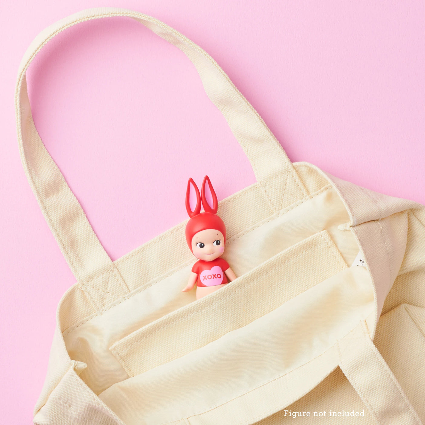 The Sonny Angel Kiss Kiss Tote Bag - Limited Edition in beige canvas features a small, red-eared Sonny Angel doll peeking from the pocket. Set against a pink background, text reads “Figure not included,” adding charm and exclusivity to your ensemble.