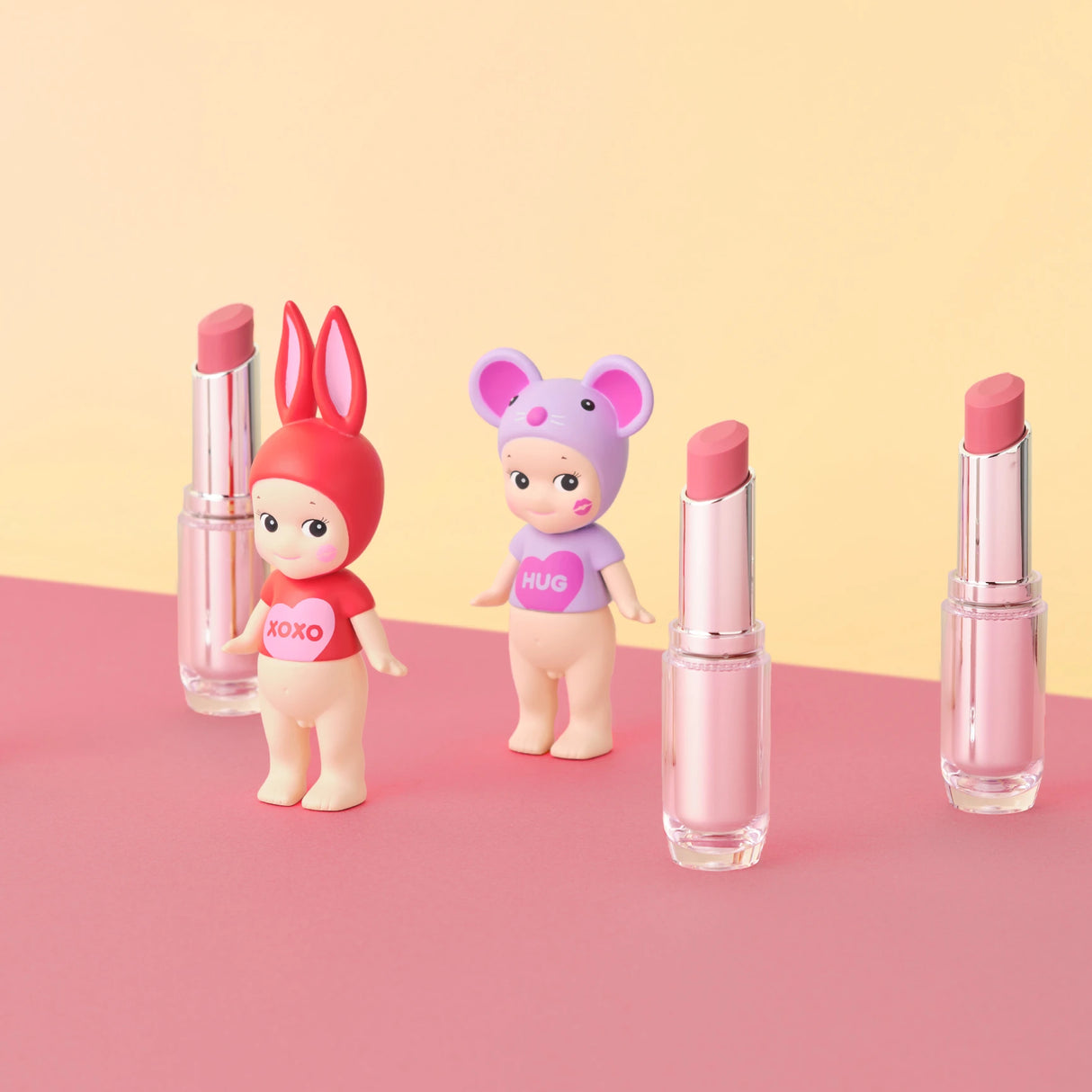 Two Sonny Angel Kiss Kiss Series figurines in themed costumes are positioned between four pink lipsticks, set against a pastel background.