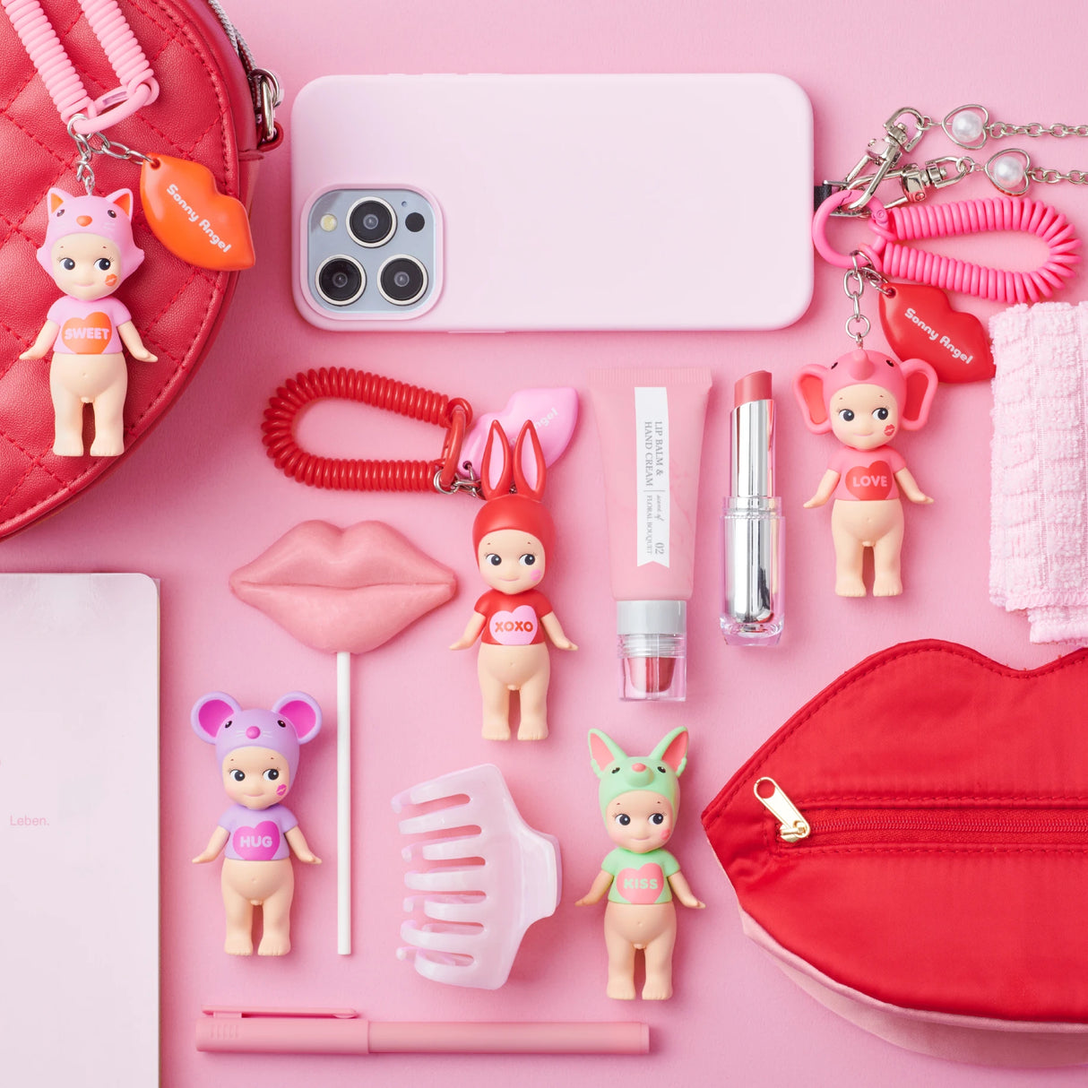 A flatlay featuring Sonny Angel Kiss Kiss Series - Blind Box dolls, makeup, a smartphone, and accessories in pink and red hues on a pink background perfectly encapsulates the charm of the collection.