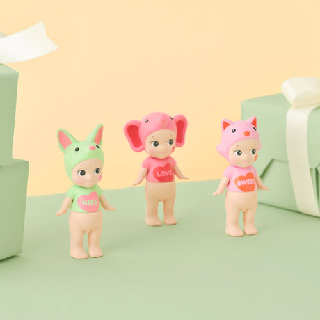 Three small Sonny Angel figures from the Kiss Kiss Series stand on a green surface with wrapped boxes. They feature animal costumes: a green bunny labeled "KISS," a pink elephant labeled "LOVE," and a pink fox labeled "SWEET.