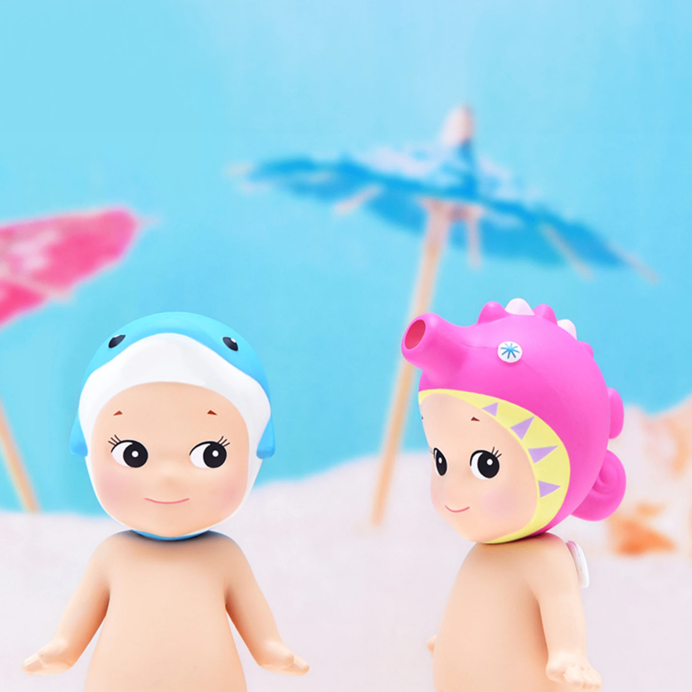 Set against a backdrop of vibrant paper umbrellas under a light blue sky, two mystery figures wearing adorable animal hats—a blue shark and a pink dinosaur—perfectly embody the charm of the Sonny Angel Marine Series by Sonny Angel, creating an ideal scene for collectors.