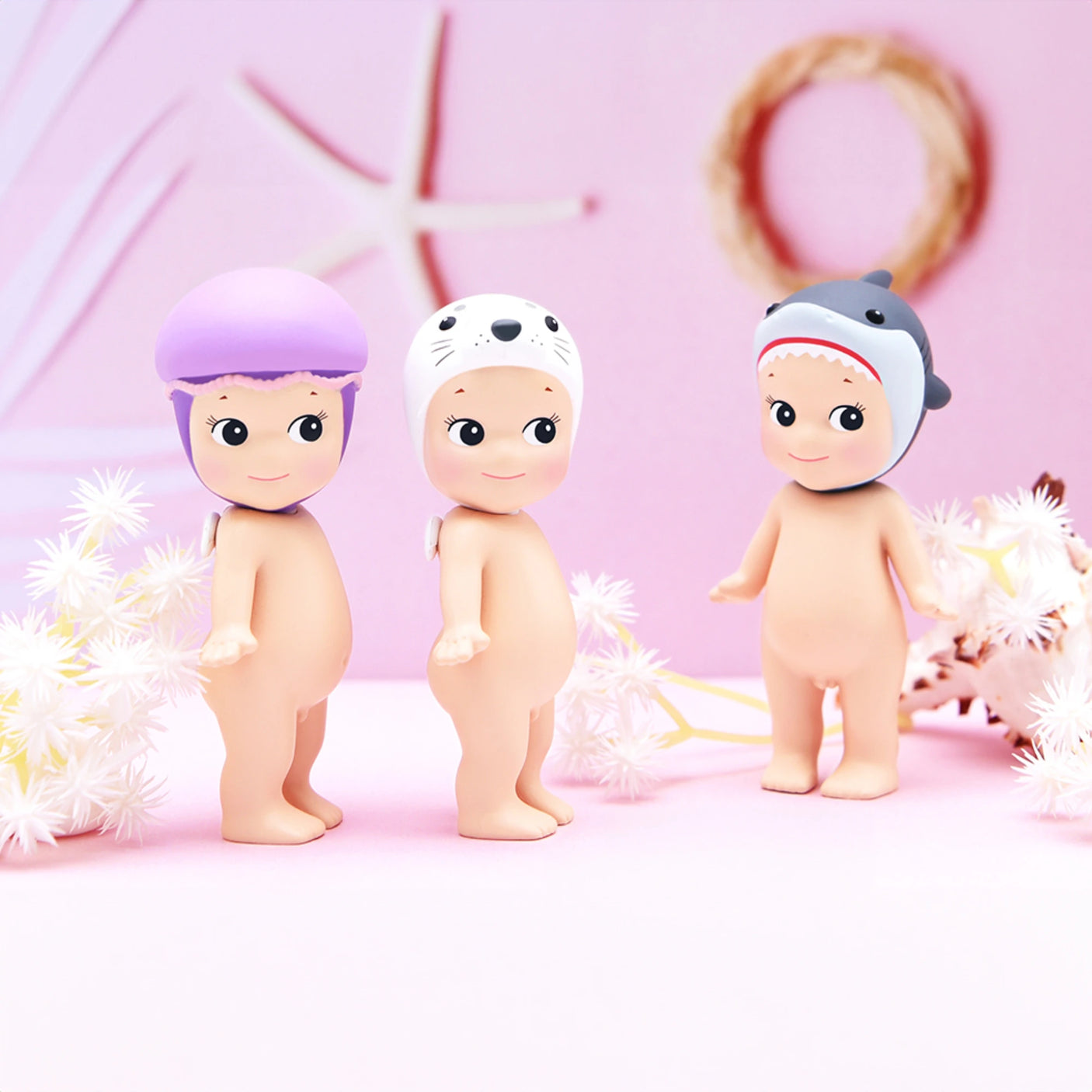 Three adorable figurines from the Sonny Angel Marine Series, each wearing an animal-themed helmet, are displayed on a pastel background adorned with starfish and floral decorations, making them a dream for every collector.