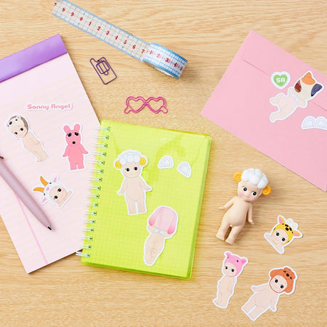 A desk with a yellow notebook, a pink notepad, a Sonny Angel Blind Box - Sticker Pack Series 1 featuring small figurines, paperclips, a measuring tape, and a pink envelope.