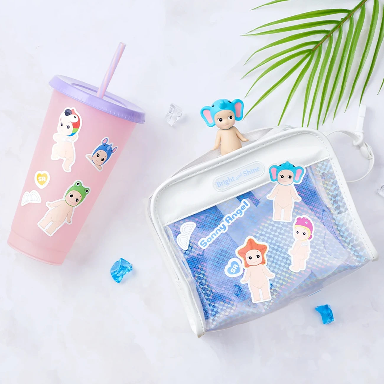 A pink tumbler decorated with water-resistant stickers is placed next to a white mesh bag featuring delightful Sonny Angel Blind Box - Sticker Pack Series 1 stickers. A small figurine with blue hair sits playfully on top of the bag, accompanied by a green leaf and blue candy pieces that enhance the whimsical atmosphere.