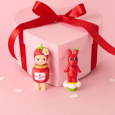 Pink heart-shaped gift box with red ribbon next to a strawberry doll and red spotted rabbit figurine on pink background