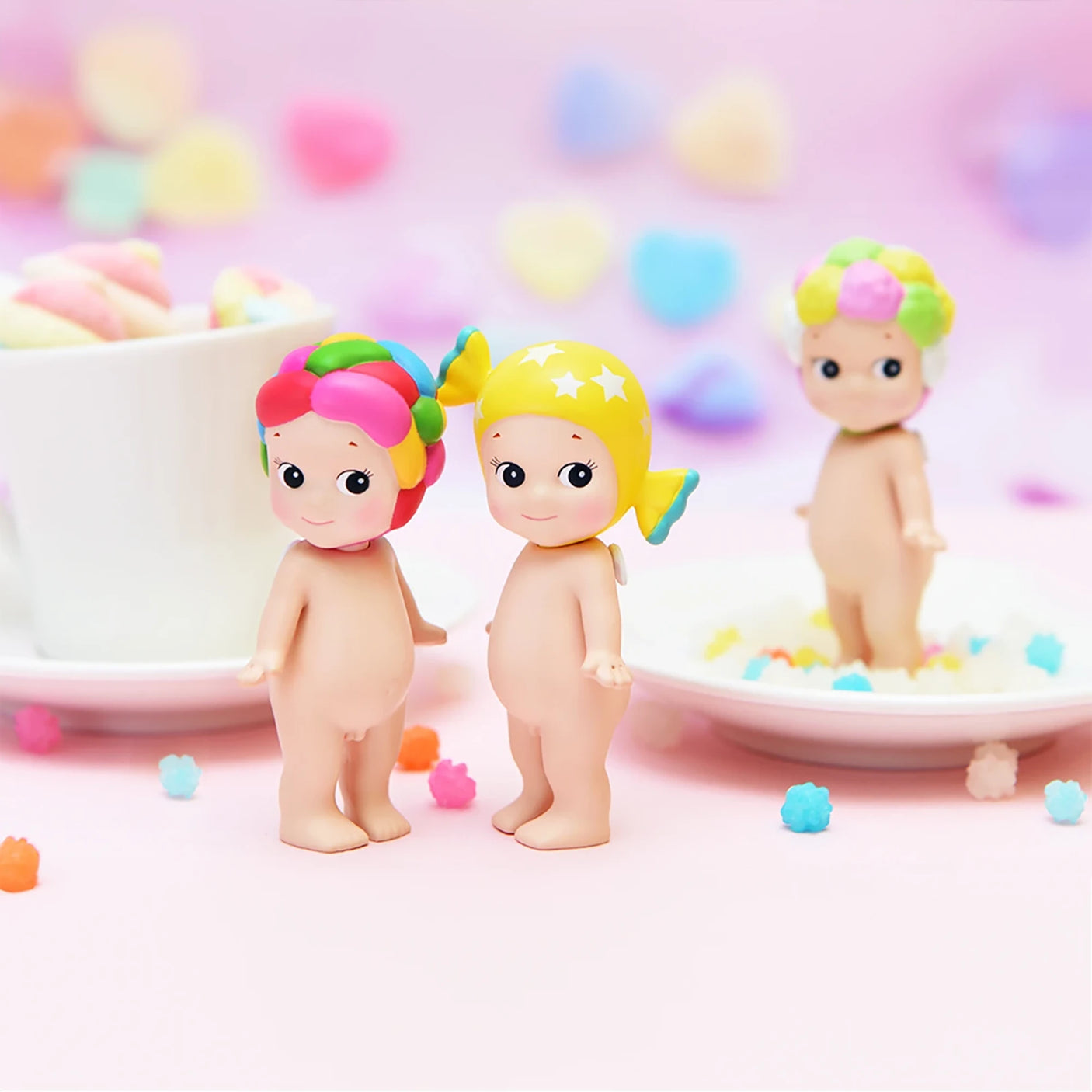 Three small, vibrant collectible figures with distinctive hairstyles are set against a pastel backdrop, evoking the charm of the Sonny Angel Sweets Series Blind Box. A cup and candies complete the whimsical scene, enticing you to discover which surprise is hidden within this delightful collection by Sonny Angel.