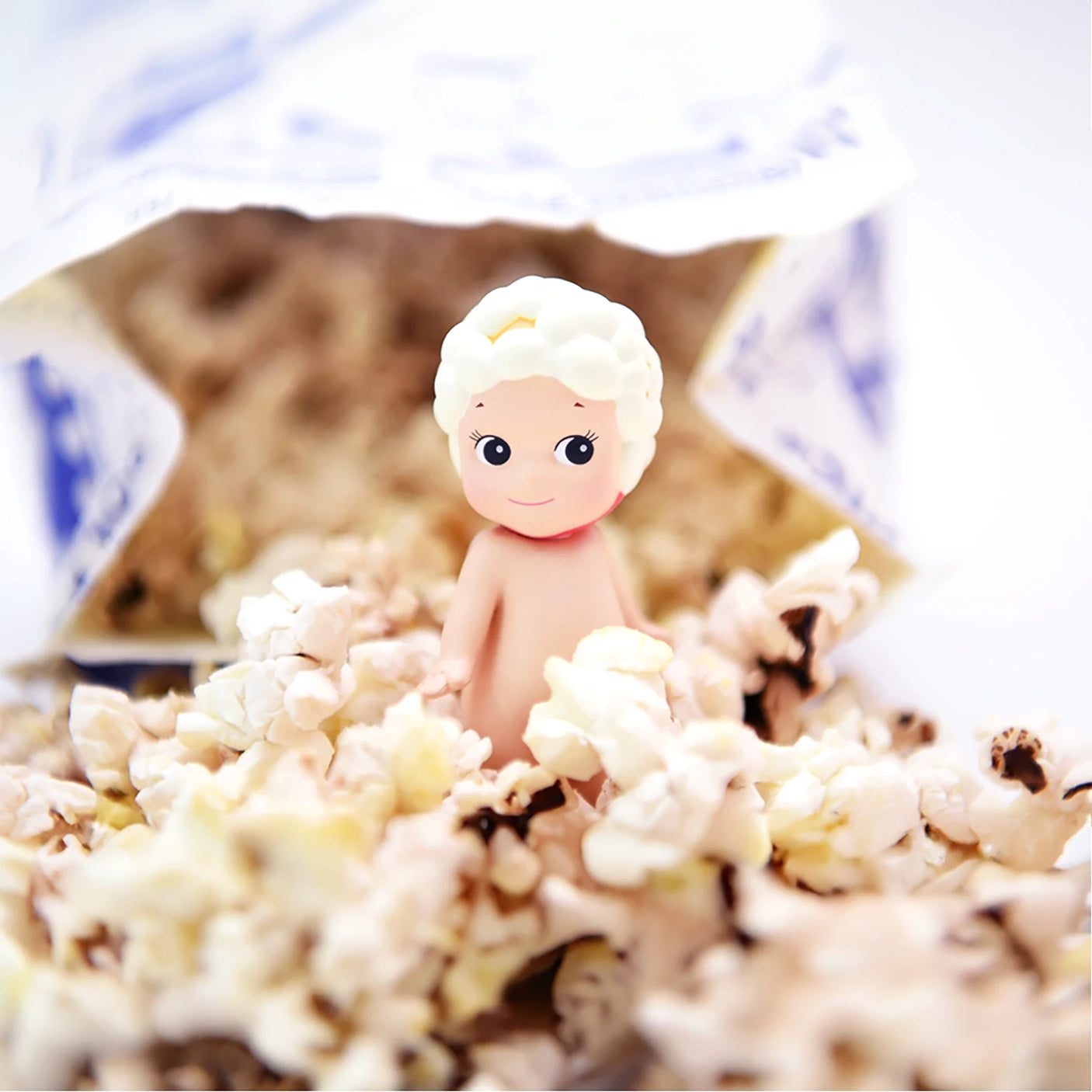 A small figurine with white curly hair is nestled in a pile of popcorn spilling out from a bag, evoking the charm of a Sonny Angel Sweets Series Blind Box by Sonny Angel.