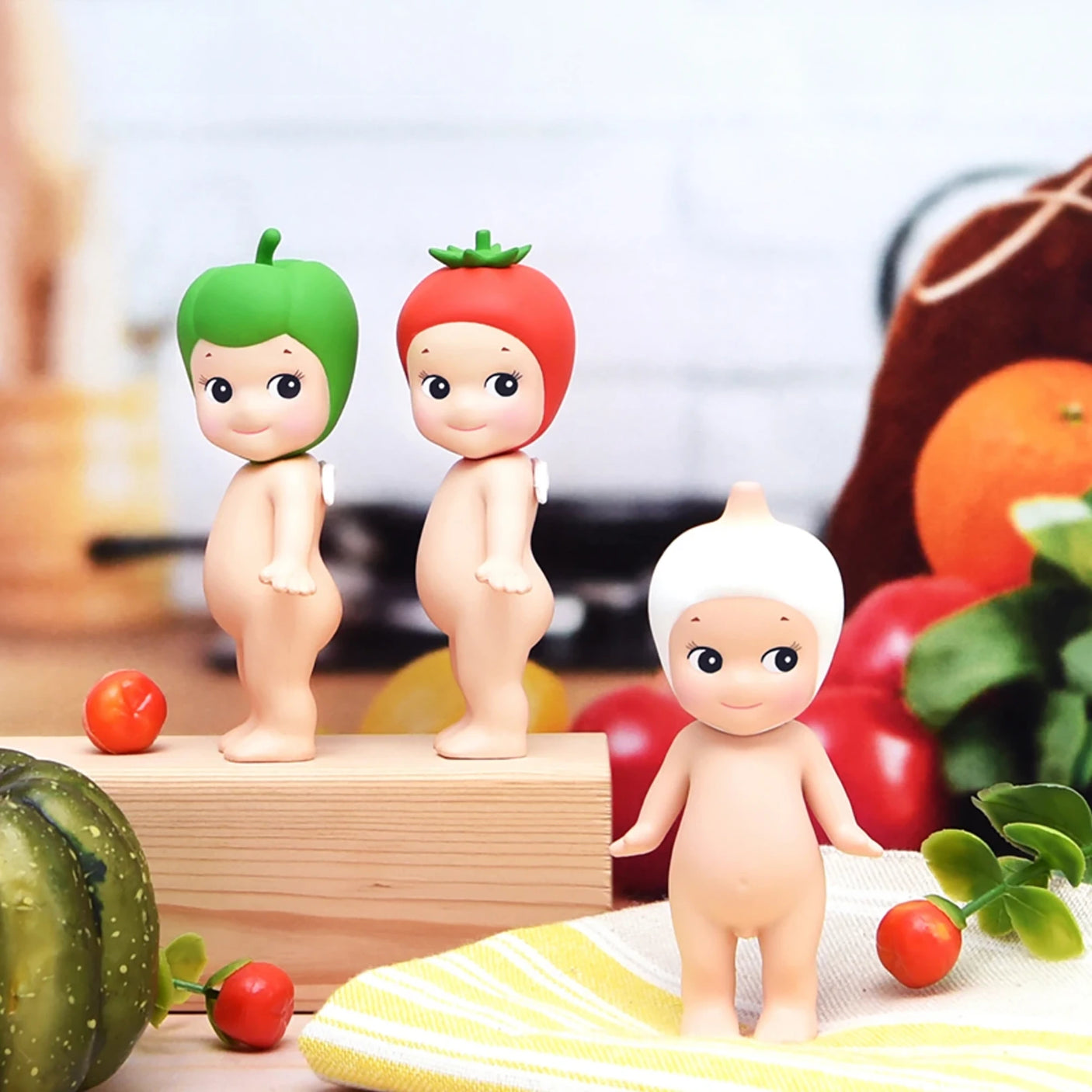 Three small figures from the Sonny Angel Vegetable Series Blind Box are displayed on a table surrounded by an assortment of fruits and vegetables.