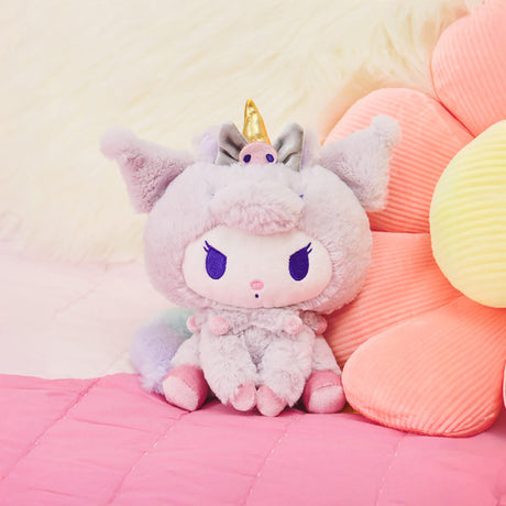 The Sanrio Kuromi Plush - Unicorn by Hello Kitty, featuring a cartoon character with purple eyes and a unicorn hoodie—ideal for Sanrio enthusiasts—is placed on a pink quilt beside an oversized pastel flower pillow.