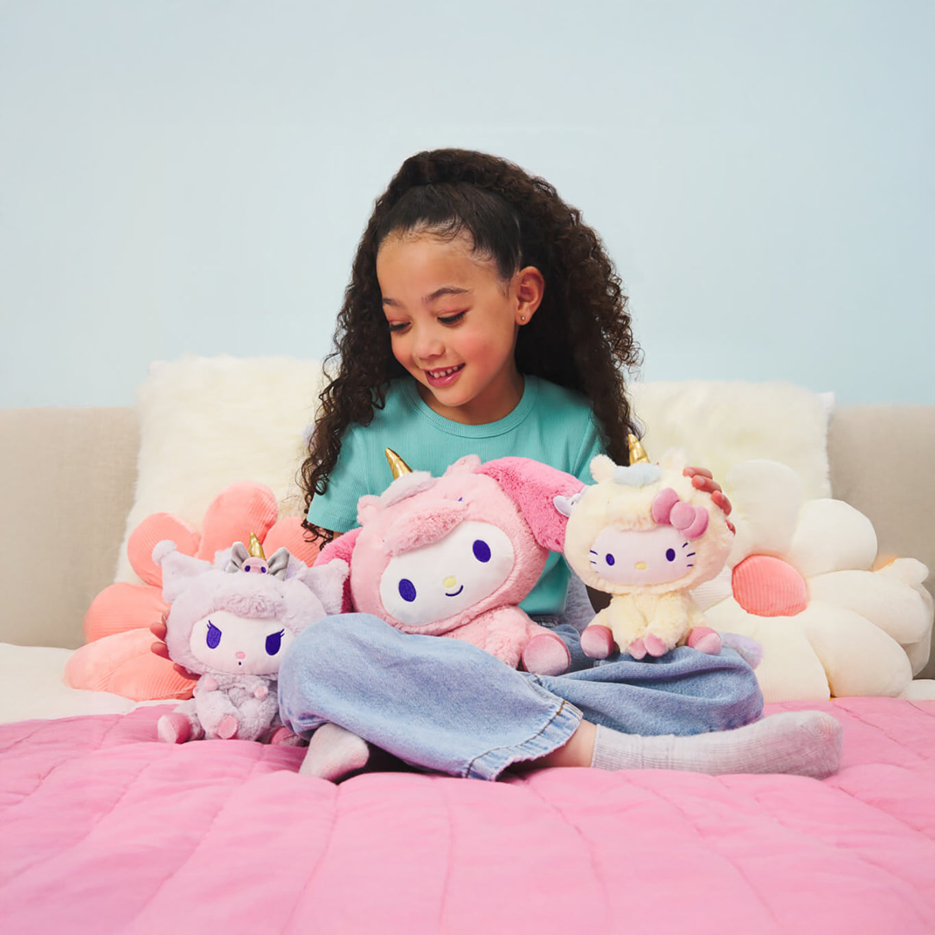 A child sits on a bed with pink covers, surrounded by three colorful plush unicorns, including the enchanting Hello Kitty Plush - Unicorn. This charming scene makes for an ideal gift for any little dreamer.