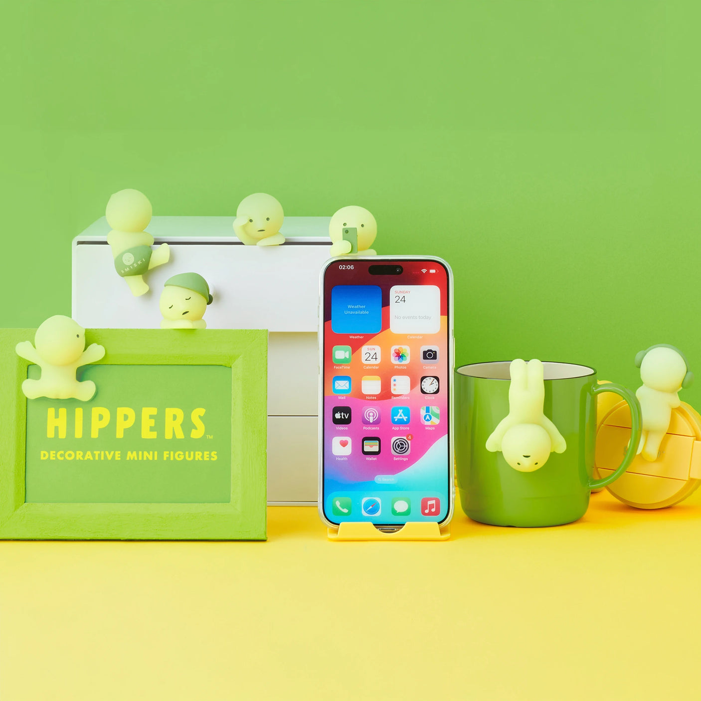 A vibrant phone is encircled by several small Smiski Hippers from their Blind Box collection, infusing a whimsical vibe with their glow-in-the-dark allure, all arranged atop boxes and a mug against a lively background.