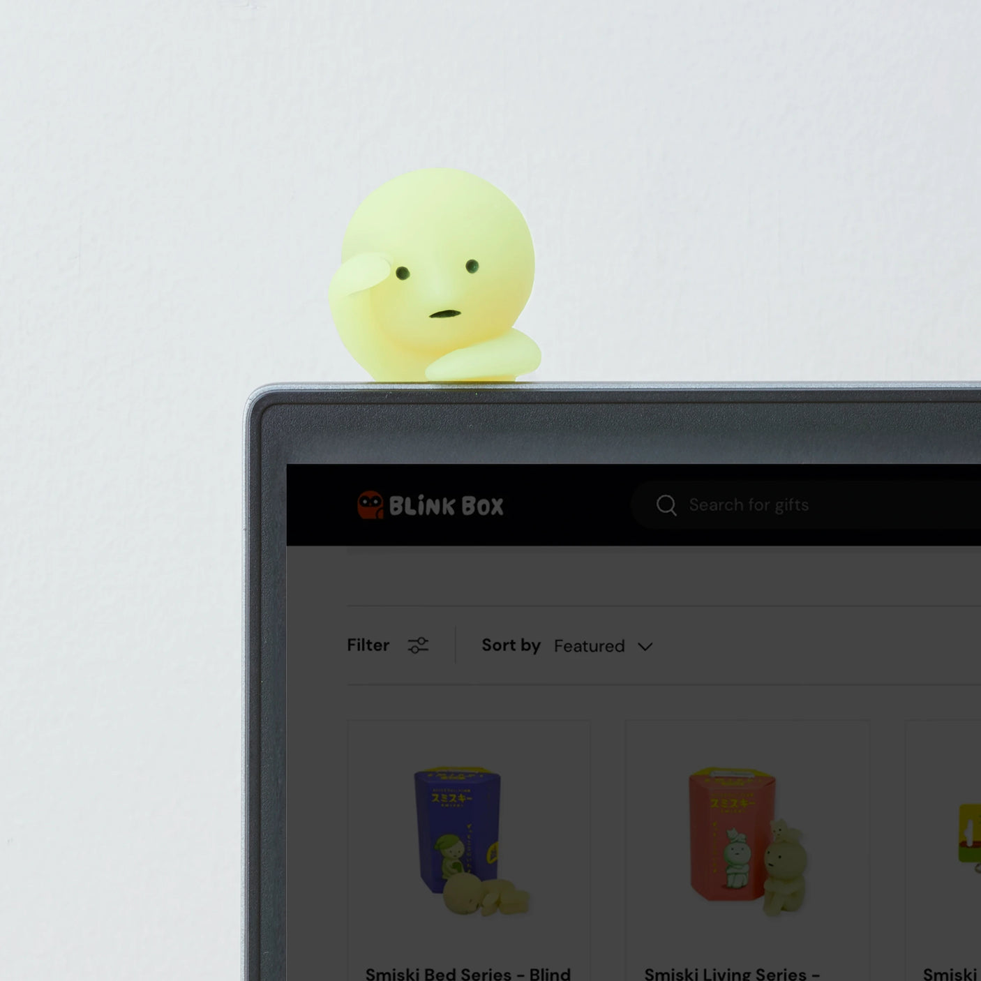 A small Smiski Hippers Blind Box figurine, glow-in-the-dark, is perched on the top corner of a monitor displaying a shopping website page.