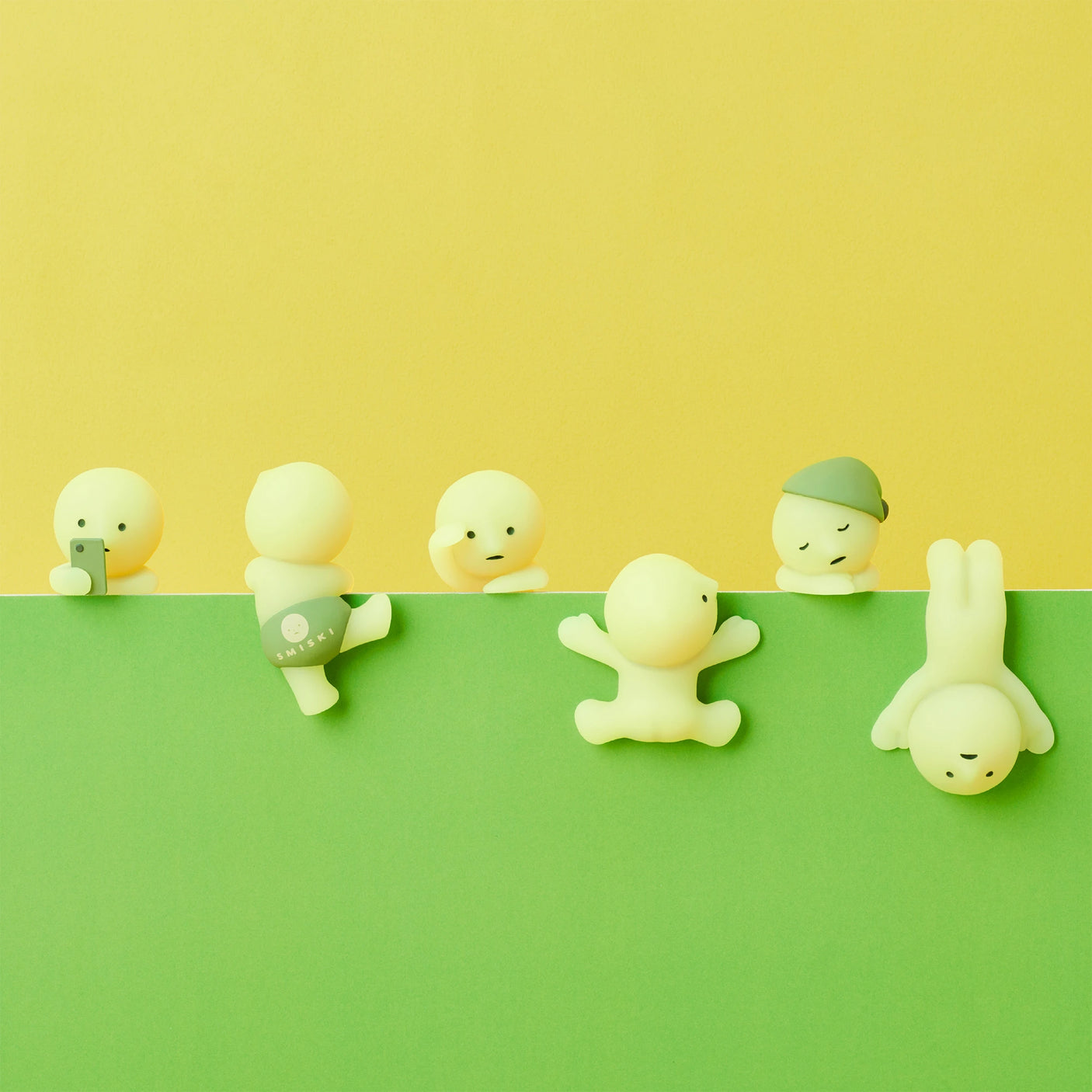 A collection of small, yellow and green humanoid figures from the Smiski Hippers Blind Box by Smiski are arranged on a matching two-tone yellow and green backdrop. Each figure showcases unique poses and expressions, with some exhibiting an enchanting glow-in-the-dark feature.