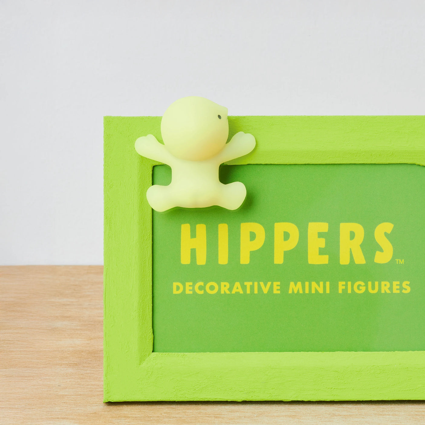 A small yellow figure on a green frame, labeled "Smiski Hippers Blind Box," sits on a wooden surface, its glow-in-the-dark charm hinting at the surprises typical of Smiski's offerings.