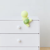 A small lime-green figurine from the Smiski Hippers Blind Box collection by Smiski rests on the edge of a white dresser. It features a green belt adorned with a logo. The dresser has three drawers with round knobs, and when the lights go out, the figure reveals its glow-in-the-dark charm.