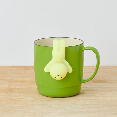 Green mug adorned with a small, yellow human-shaped figure from the Smiski Hippers Blind Box collection, hanging upside down from the rim on a wooden surface.