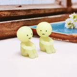 Two small figurines from the Smiski Series 1 - Blind Box by Smiski, featuring simple facial expressions and a yellow hue, sit on a table near a wooden window frame and a blue book. They cast an enchanting charm as they subtly glow-in-the-dark.