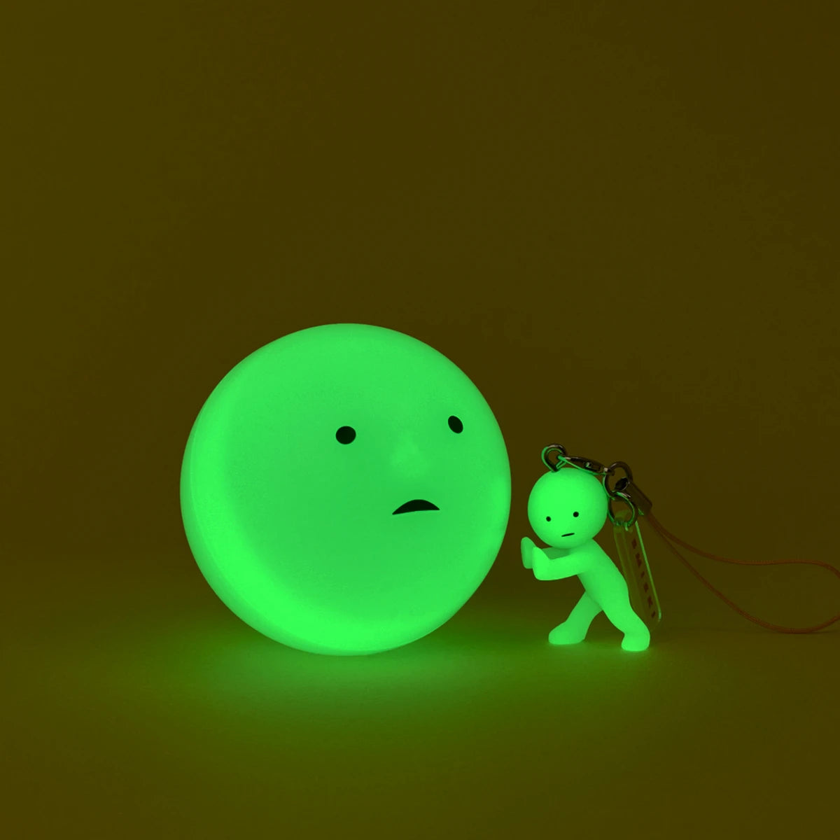 The Smiski Strap Accessory Keychain - Series 2 by Smiski features a small, glowing green humanoid figurine pushing a larger glowing ball with a simple face—ideal as a glow-in-the-dark keychain for collectible mini figure fans.