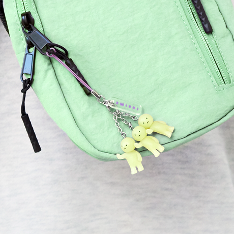 Three small pale yellow character charms with simple faces hanging from a purple strap on light green bag zipper
