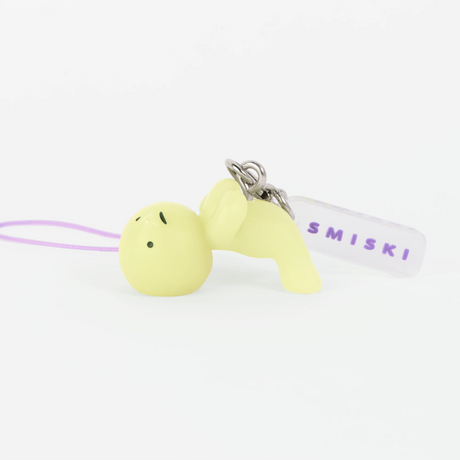 Small pale yellow character keychain lying on its back with purple strap and clear tag on white background
