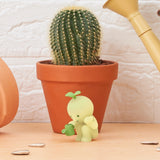 A cactus in a terracotta pot sits behind a small green Smiski Sunday Series figure holding a watering can. Scattered seeds shimmer on the wooden surface, while the glow-in-the-dark Smiski adds an enchanting touch to the scene.