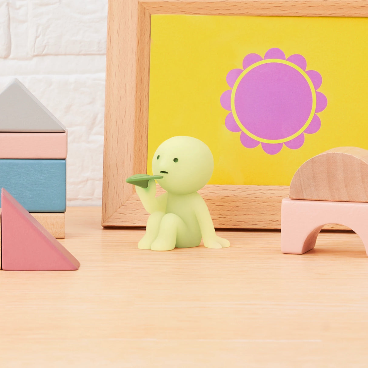 A small glow-in-the-dark figure from the Smiski Sunday Series - Blind Box stands with a leaf on wood near blocks and a framed yellow print featuring pink and purple elements.