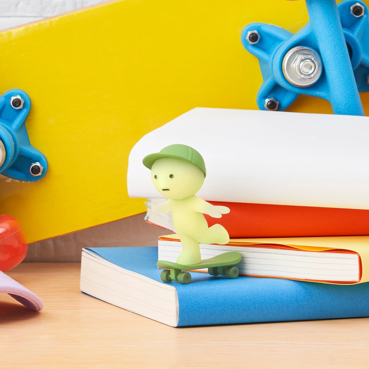 A small green Smiski figurine with a cap rides a skateboard atop colorful books from the Smiski Sunday Series - Blind Box. It glows in the dark, and skateboard wheels plus a toy add to this playful scene.