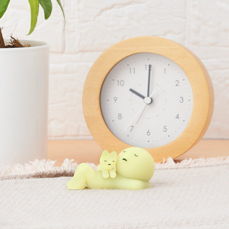 A small, glow-in-the-dark figure from the Smiski Sunday Series and a cat relax on a beige mat. A round wooden clock and a white plant pot complete the cozy scene, embodying the charm of Smiski.