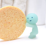 A glow-in-the-dark collectible from the Smiski Bath Series - Blind Box by Smiski shows what looks like a small blue figurine playfully pushing against a large, round, textured sponge on a light background, adding whimsical charm to any bathroom setting.