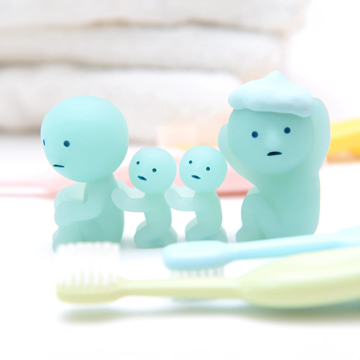 Four figures from the Smiski Bath Series by Smiski, featuring light blue round-headed figurines with small bodies, are lined up and holding each other. The largest figure stands out with a piece of white material on its head, adding a unique touch to the collection. In front of these collectible figures are three toothbrushes.