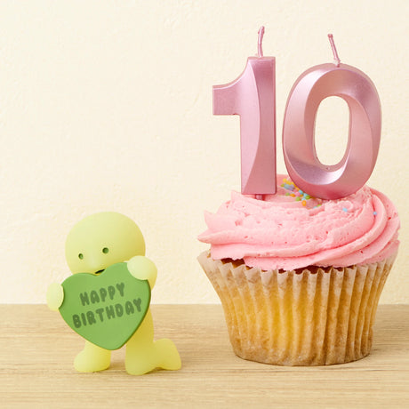 A pink frosted cupcake with pink number 10 candles sits beside a yellow Smiski Birthday Series - Blind Box figure from Smiski holding a green heart-shaped "Happy Birthday" sign—a delightful glow-in-the-dark surprise.