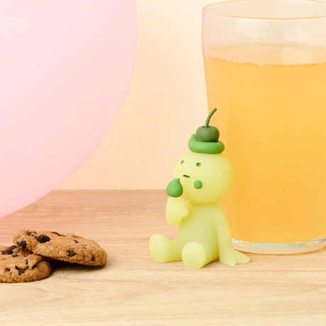 A small green Smiski Birthday Series figurine from Smiski sits on a table with a glass of orange juice, two chocolate chip cookies, and a pink balloon.