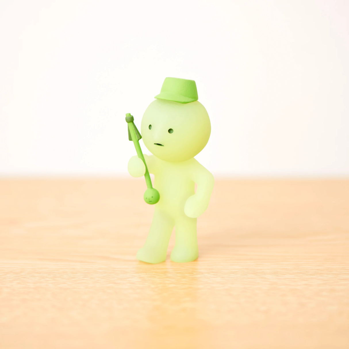 A charming miniature from the Smiski Cheer Series, featuring a small green figure with a hat and staff on a light wooden base, is part of the delightful collection of glow-in-the-dark figures available in each blind box by Smiski.